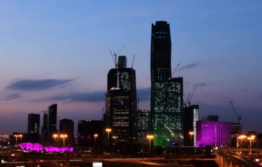 City skyline at dusk featuring tall skyscrapers with construction cranes, illuminated windows, and vibrant purple and green lights at street level—highlighting achievements inspired by the Ministry of Energy KSA as evening skies shift from blue to purple.